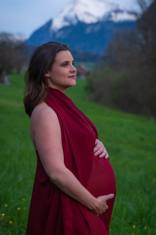 Schwangere Frau in rotem Kleid steht vor Bergkulisse